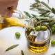 "Olio extra vergine di oliva versato da una caraffa in un bicchiere, con rami di ulivo e olive verdi fresche su sfondo chiaro.