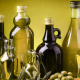 Diverse bottiglie di olio extravergine di oliva, di varie forme e colori, disposte su un tavolo con rami di ulivo e olive verdi in primo piano.