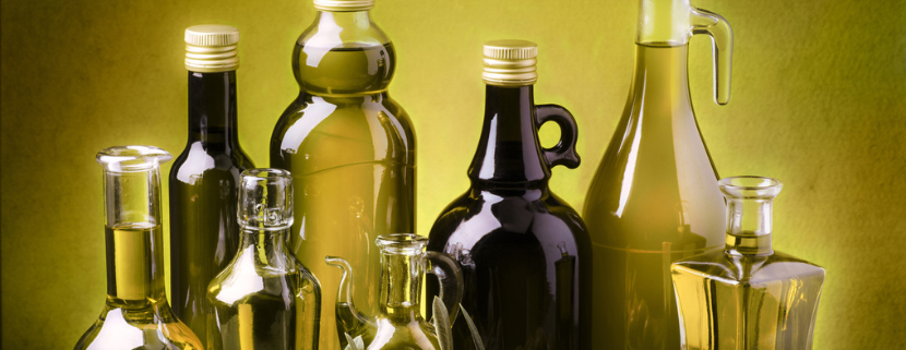 Diverse bottiglie di olio extravergine di oliva, di varie forme e colori, disposte su un tavolo con rami di ulivo e olive verdi in primo piano.