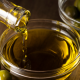 Olio extravergine di oliva versato in una ciotola di vetro con olive verdi accanto, su sfondo in legno scuro.