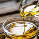 Olio extravergine di oliva versato da una caraffa in una ciotola di vetro su fondo rustico in legno, con ramoscelli d’ulivo decorativi
