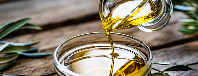 Olio extravergine di oliva versato da una caraffa in una ciotola di vetro su fondo rustico in legno, con ramoscelli d’ulivo decorativi