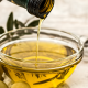 Olio extravergine di oliva versato in una ciotola di vetro con olive verdi e utensili da cucina sullo sfondo.