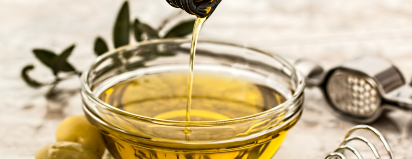 Olio extravergine di oliva versato in una ciotola di vetro con olive verdi e utensili da cucina sullo sfondo.