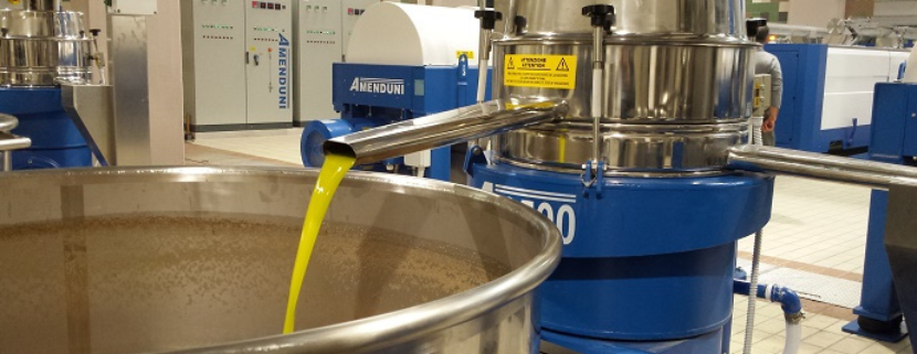 Frantoio oleario industriale durante il processo di estrazione dell’olio extravergine di oliva, con macchinari in acciaio inox e flusso di olio in uscita in primo piano.