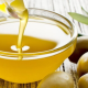 Ciotola di vetro con olio extravergine di oliva dorato, circondata da olive verdi e foglie di ulivo su sfondo in legno chiaro.
