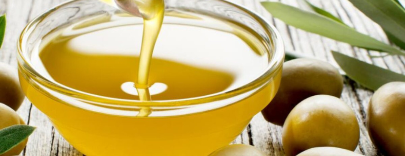Ciotola di vetro con olio extravergine di oliva dorato, circondata da olive verdi e foglie di ulivo su sfondo in legno chiaro.