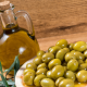 Bottiglia di vetro con olio extravergine di oliva accanto a un piatto di olive verdi su tavolo in legno, con ramoscello di ulivo decorativo.
