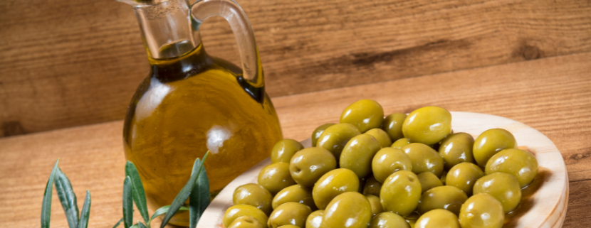 Bottiglia di vetro con olio extravergine di oliva accanto a un piatto di olive verdi su tavolo in legno, con ramoscello di ulivo decorativo.