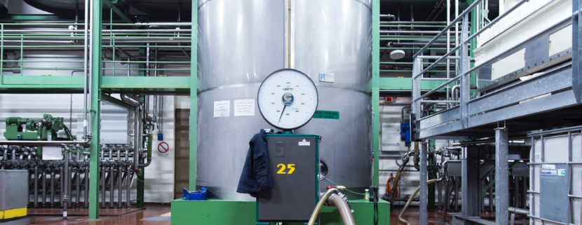 Interno di un impianto industriale per la conservazione dell'olio extravergine di oliva, con grandi serbatoi in acciaio inox, manometri, tubazioni e strutture metalliche per il controllo della qualità e dello stoccaggio del prodotto.