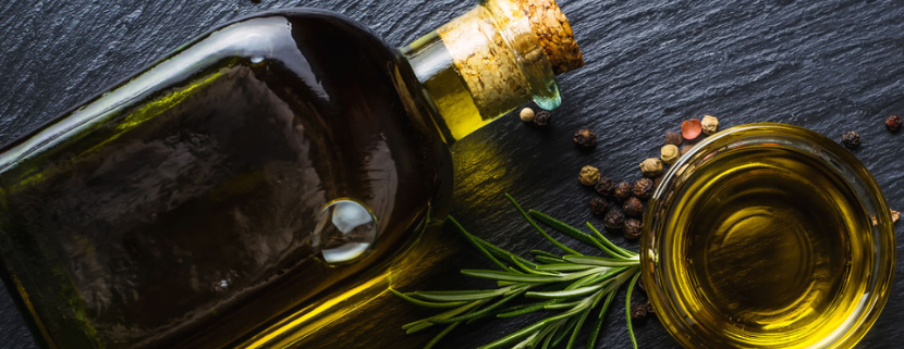 Bottiglia e ciotola di olio extravergine d'oliva su sfondo scuro, accompagnate da rametti di rosmarino e grani di pepe.