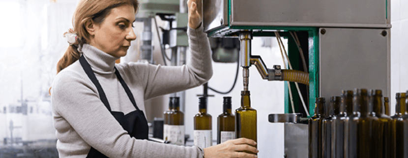 Donna che lavora in un frantoio imbottigliando olio extravergine di oliva in bottiglie di vetro scuro tramite una macchina automatica per l’imbottigliamento.