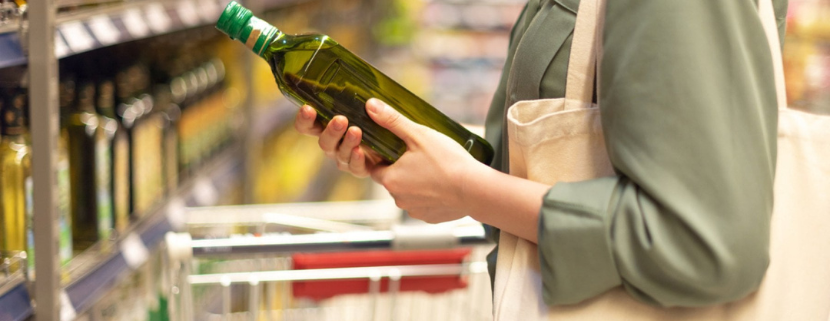 Donna con borsa di tela osserva attentamente una bottiglia di olio extravergine di oliva in corsia supermercato, accanto a uno scaffale pieno di prodotti simili e un carrello della spesa.