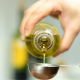 Mano che versa olio extravergine di oliva da una bottiglia in un cucchiaio dosatore in acciaio, con sfondo sfocato da cucina.