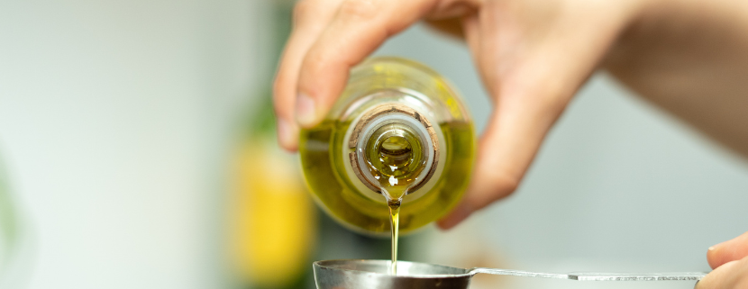 Mano che versa olio extravergine di oliva da una bottiglia in un cucchiaio dosatore in acciaio, con sfondo sfocato da cucina.