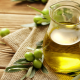 Due caraffe di vetro piene di olio extravergine di oliva su un tavolo rustico in legno, circondate da rami di ulivo e olive verdi fresche, simbolo di tradizione, genuinità e produzione artigianale.