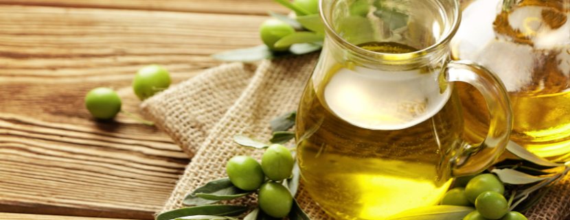 Due caraffe di vetro piene di olio extravergine di oliva su un tavolo rustico in legno, circondate da rami di ulivo e olive verdi fresche, simbolo di tradizione, genuinità e produzione artigianale.