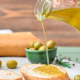 Olio extravergine di oliva versato su una fetta di pane, con olive verdi sullo sfondo, simbolo di qualità artigianale e tracciabilità nella filiera certificata.