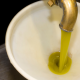 Getto di olio extravergine di oliva appena estratto che scorre da un rubinetto metallico in un contenitore bianco durante la fase di produzione in frantoio.