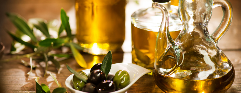 Bottiglie e caraffa di olio extravergine di oliva su tavolo in legno, con ciotola di olive fresche e rami di ulivo decorativi.