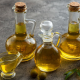 Bottiglie e oliere di vetro piene di olio d’oliva disposte su un tavolo, con alcune olive verdi in primo piano, simbolo della produzione e classificazione dell’olio di oliva.