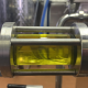 Dettaglio di un impianto di frantoio con tubo in acciaio inox che mostra il flusso di olio d’oliva appena estratto, di colore verde intenso.
