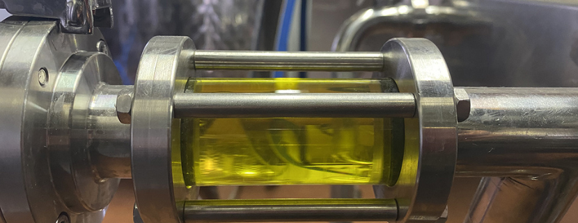 Dettaglio di un impianto di frantoio con tubo in acciaio inox che mostra il flusso di olio d’oliva appena estratto, di colore verde intenso.