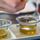 Degustazione di olio d’oliva con piccoli bicchieri trasparenti disposti su un vassoio, mentre una mano prepara l’assaggio per valutare qualità e acidità dell’olio.