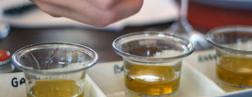 Degustazione di olio d’oliva con piccoli bicchieri trasparenti disposti su un vassoio, mentre una mano prepara l’assaggio per valutare qualità e acidità dell’olio.