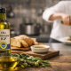 Due bottiglie di olio extravergine di oliva DOP e IGP su un tavolo in legno con olive, pane e una ciotola di olio, mentre uno chef sullo sfondo versa olio su un piatto in una cucina professionale.